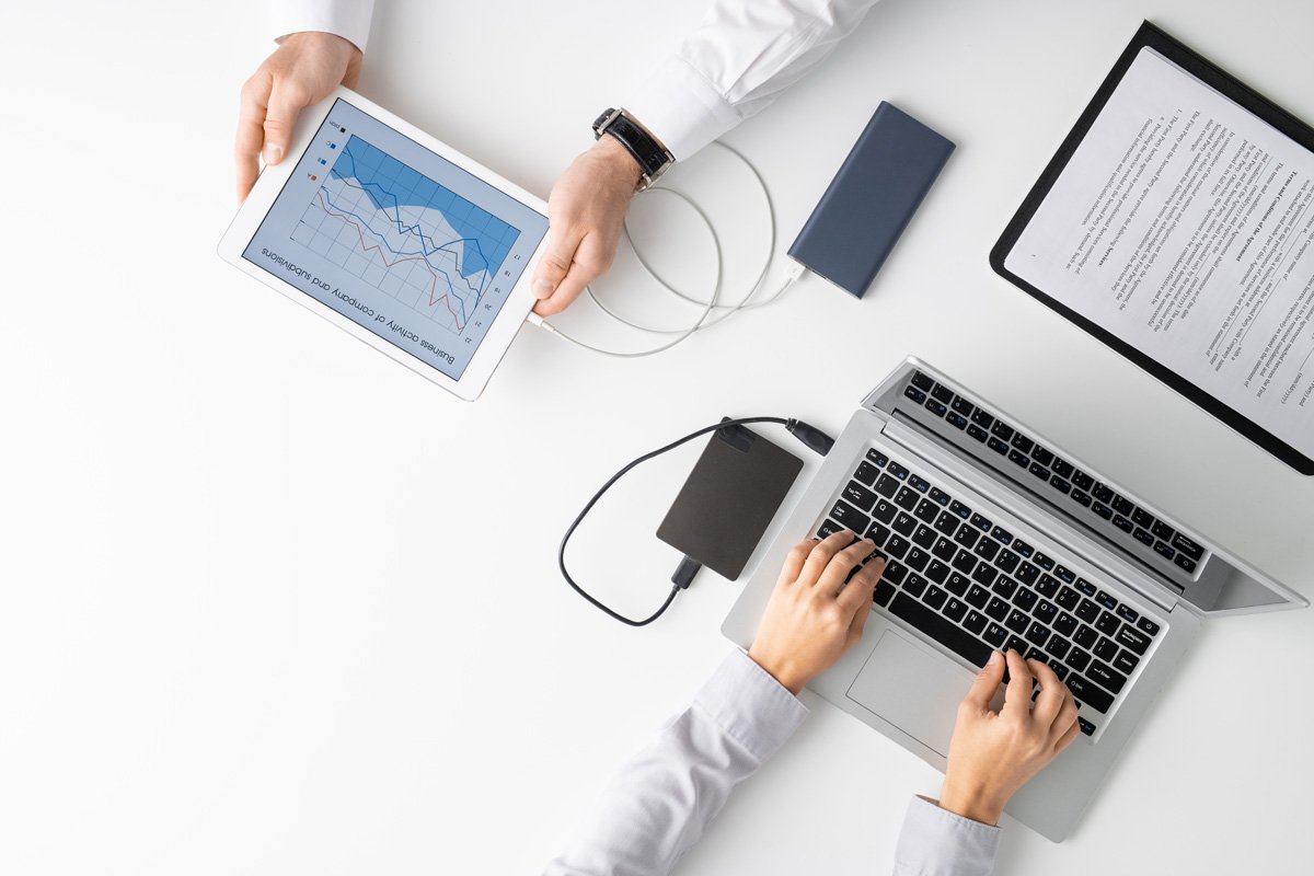 Overhead view of two people at a white desk, one holding a tablet showing a business activity chart and the other typing on a laptop